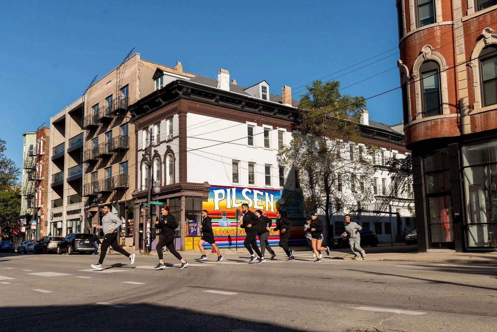 A group of runners crossing a street in Pilsen, Chicago, past a bright mural that spells out Pilsen.