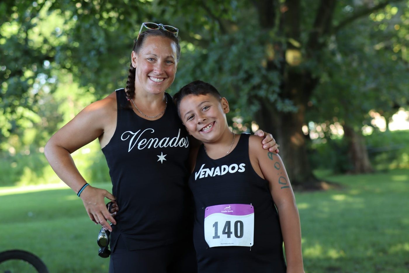 A smiling young runner wearing race bib 140 stands arm in arm with an adult runner in a park.